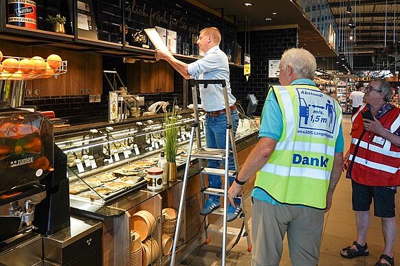 Über der Salatbar, gleich links vom Eingang: EDEKA-Marktleiter André Fiolek positioniert das "Goldene Pedal" und die Urkunde bei den anderen Auszeichnungen, unterstützt von Dr. Reinhard Spörer (gelbe ＡＤＦＣ-Weste) und Reinhard Wolf (rote ＡＤＦＣ-Weste)