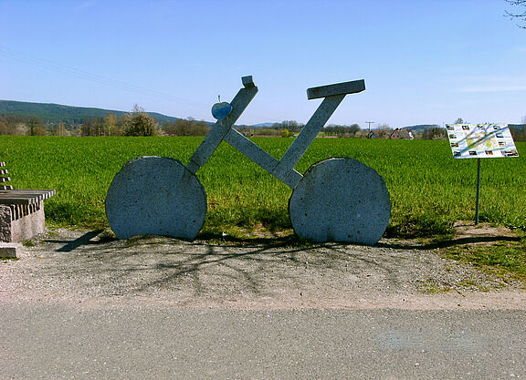 Ein Fahrrad aus Stein übergroß am Wegesrand. Dahinter eine große grüne Landschaft mit Wald am Bildrand.