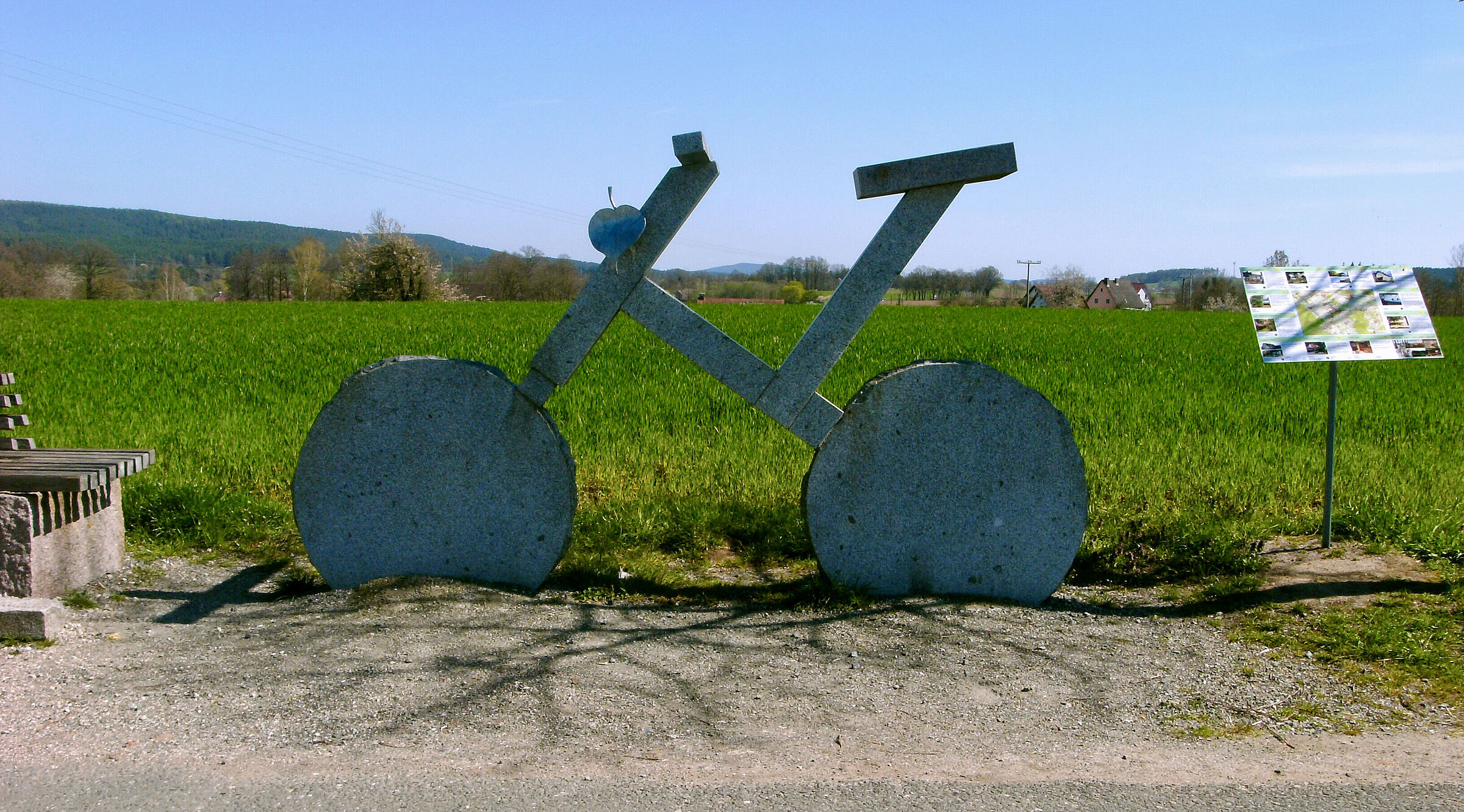 Ein übergroßes Fahrrad aus Stein steht am Wegesrand vor einer grünen Wiese.