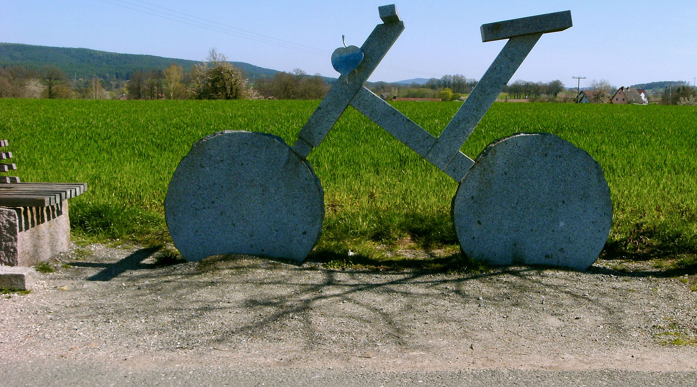 Sehr großes Fahrrad aus Stein am Wegesrand. Im Hintergrund große grüne Weide.