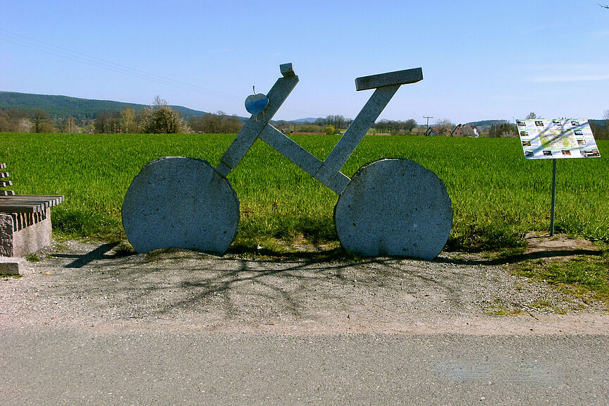 Ein übergroßes Fahrrad aus Stein steht am Wegesrand vor einer grünen Wiese.