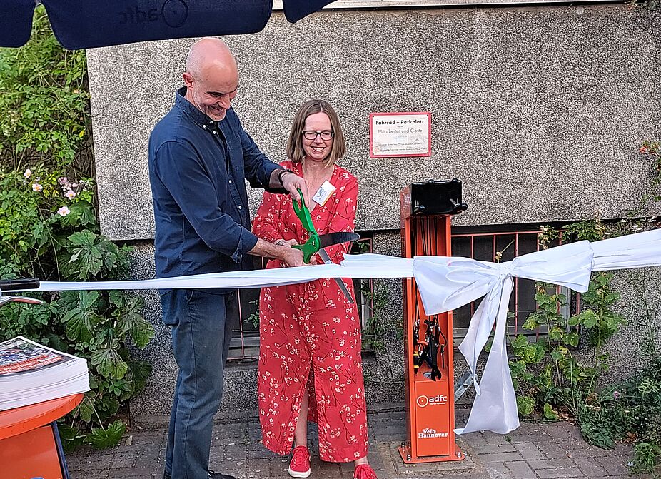 Oberbürgermeister Onay und ADFC-Vorständin Angelika Licht durchschneiden mit großer Schere das Schleifenband zur Einweihung der Fahrradreparatursäule vorm Umweltzentrum Hannover.