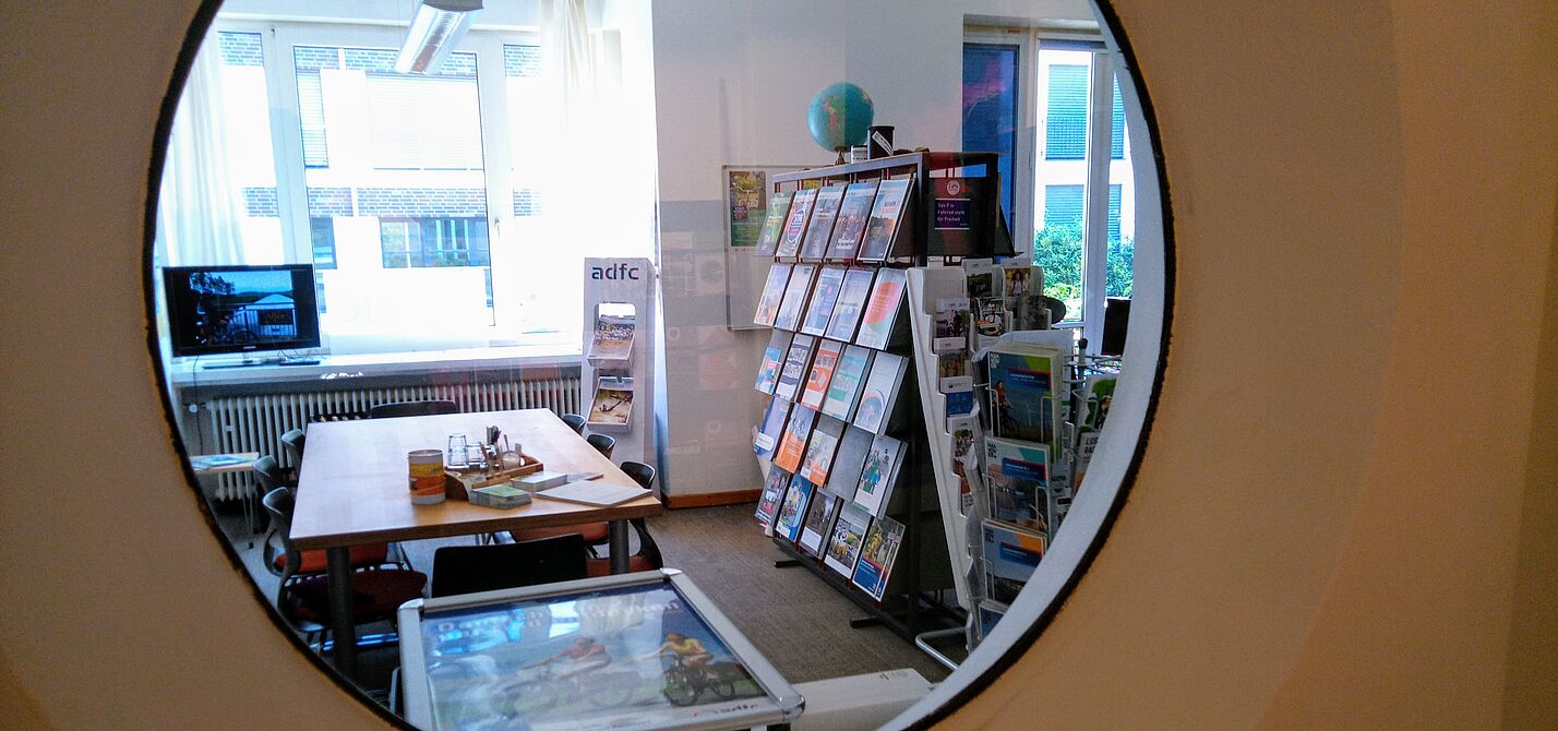 Blick durch ein Rundfenster in den ADFC-Infoladen. Es sind Regale mit Informationsmaterial, Beratungstisch und TV mit Fahrradurlaubsbildern zu sehen.