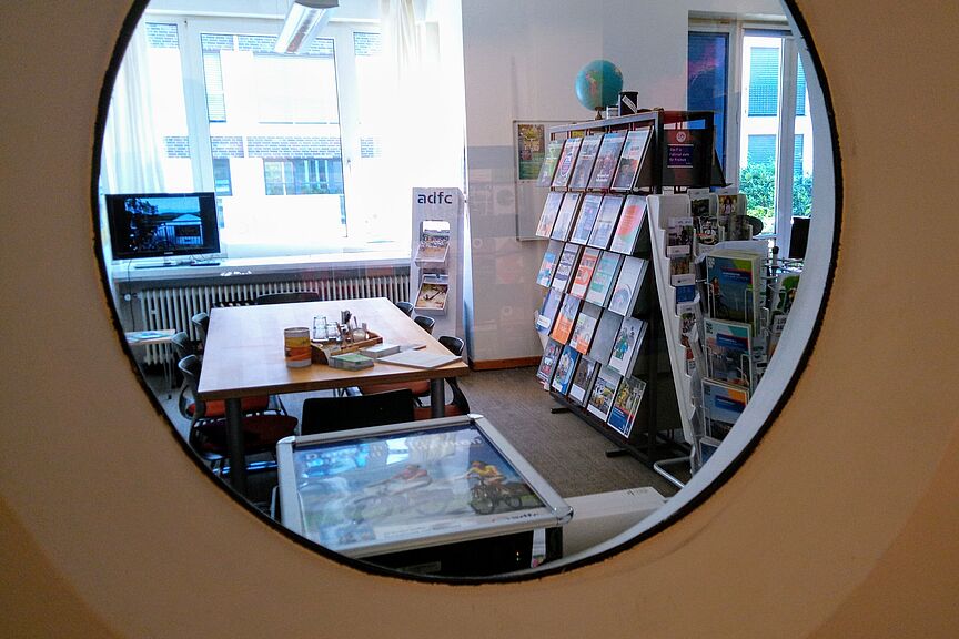 Blick durch ein Rundfenster in den ADFC-Infoladen. Es sind Regale mit Informationsmaterial, Beratungstisch und TV mit Fahrradurlaubsbildern zu sehen.