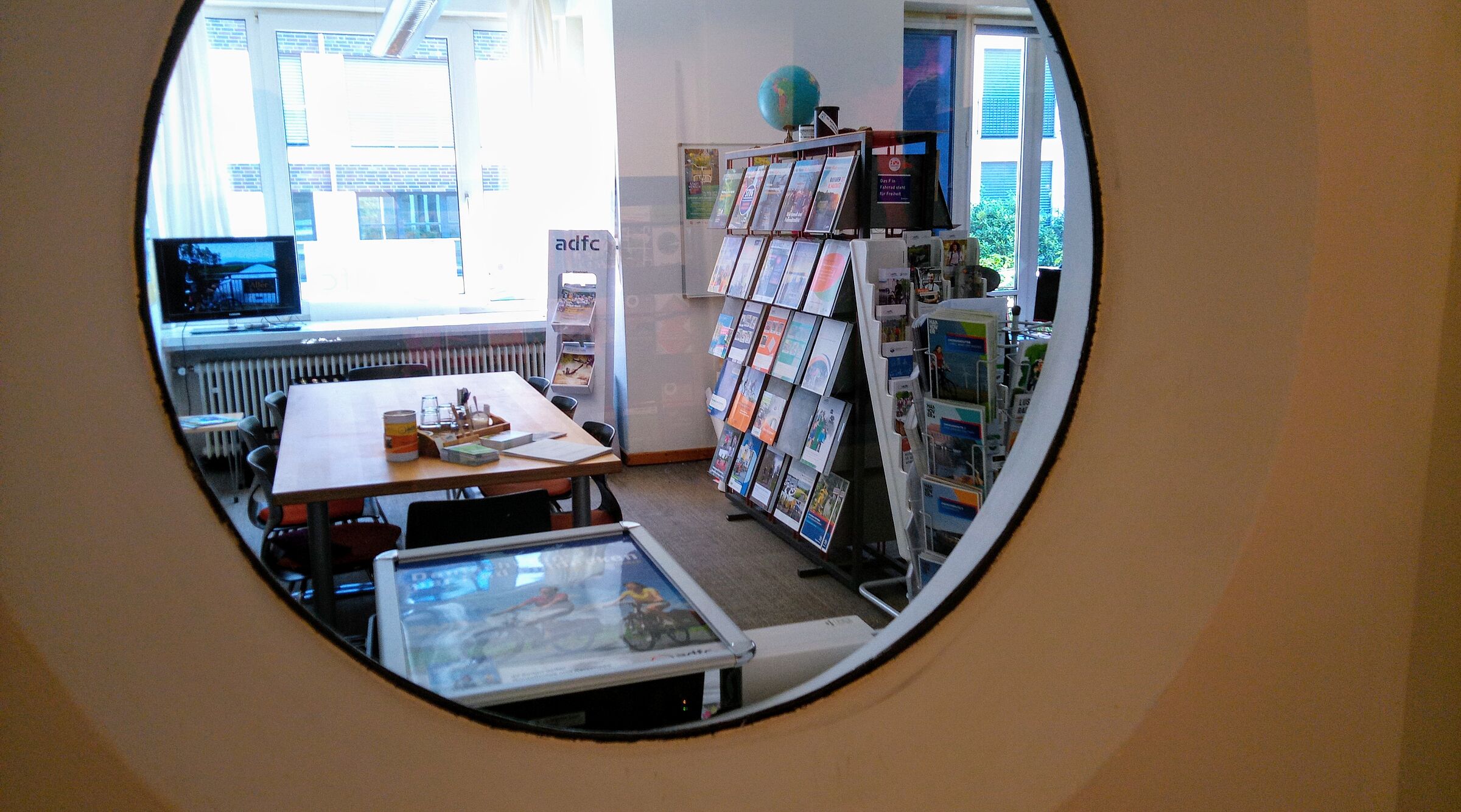 Blick durch ein Rundfenster in den ADFC-Infoladen. Es sind Regale mit Informationsmaterial, Beratungstisch und TV mit Fahrradurlaubsbildern zu sehen.
