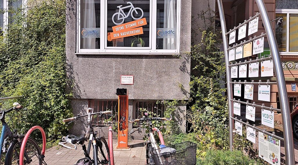 Hinter roten Fahrradbügeln steht eine orangefarbende Fahrradreparatursäule. Darüber ist das Fenster der ADFC-Geschäftsstelle zu sehen mit der Beschriftung "Deine Stimme für den Radverkehr" unter einem weißem Fahrrad.