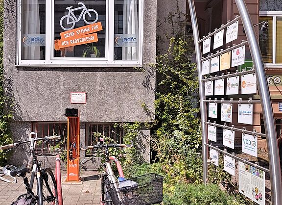 Hinter roten Fahrradbügeln steht eine orangefarbende Fahrradreparatursäule. Darüber ist das Fenster der ADFC-Geschäftsstelle zu sehen mit der Beschriftung "Deine Stimme für den Radverkehr" unter einem weißem Fahrrad.