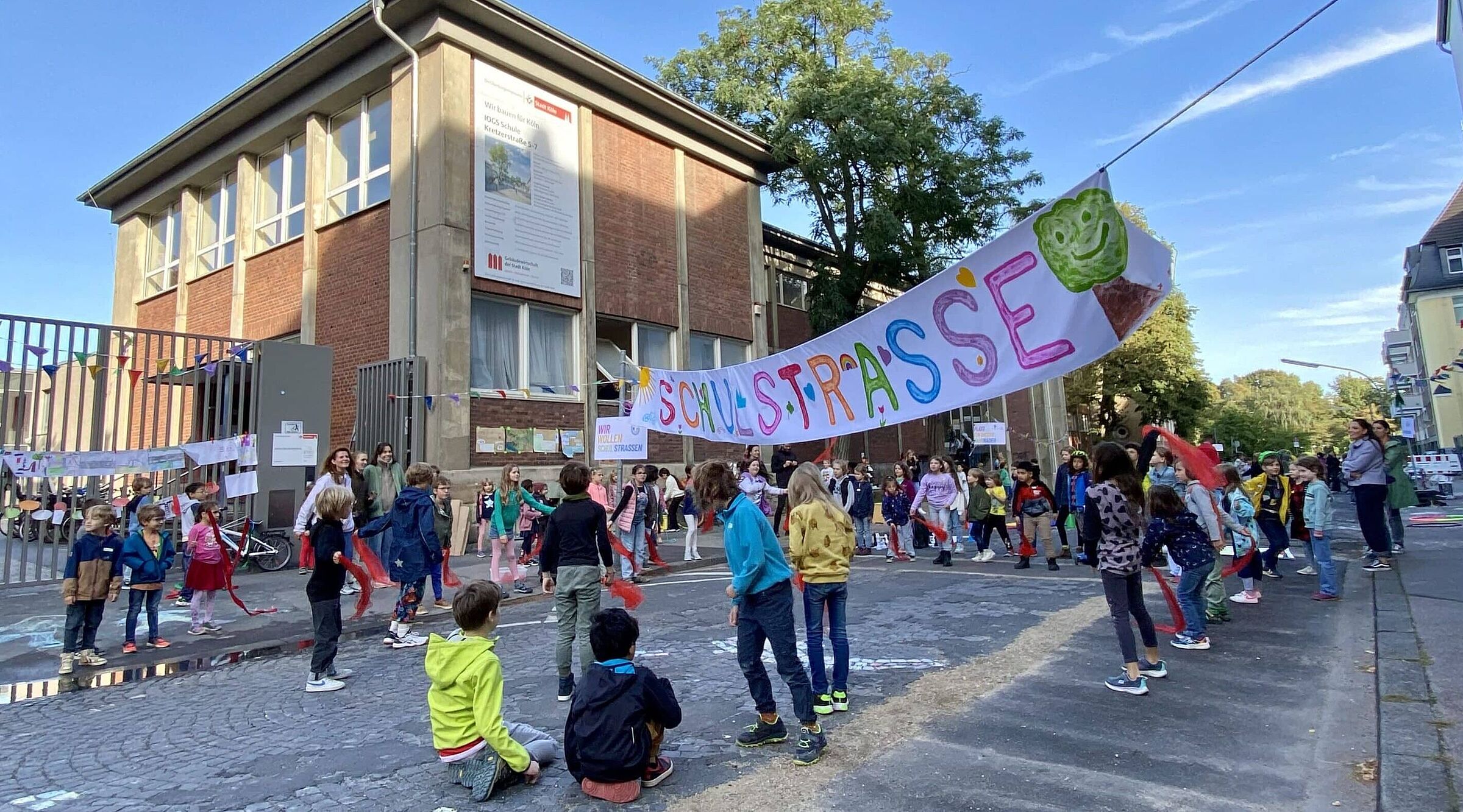 Kinder verschiedenen Alters spielen und bewegen sich auf einer gepflasterten Straße vor einem zweistöckigen Backsteingebäude. Über der Straße hängt ein buntes Banner mit der Aufschrift „Schulstraße“ und einem grünen Baum-Symbol. Im Hintergrund sind Erwachsene zu sehen, die die Aktion begleiten.