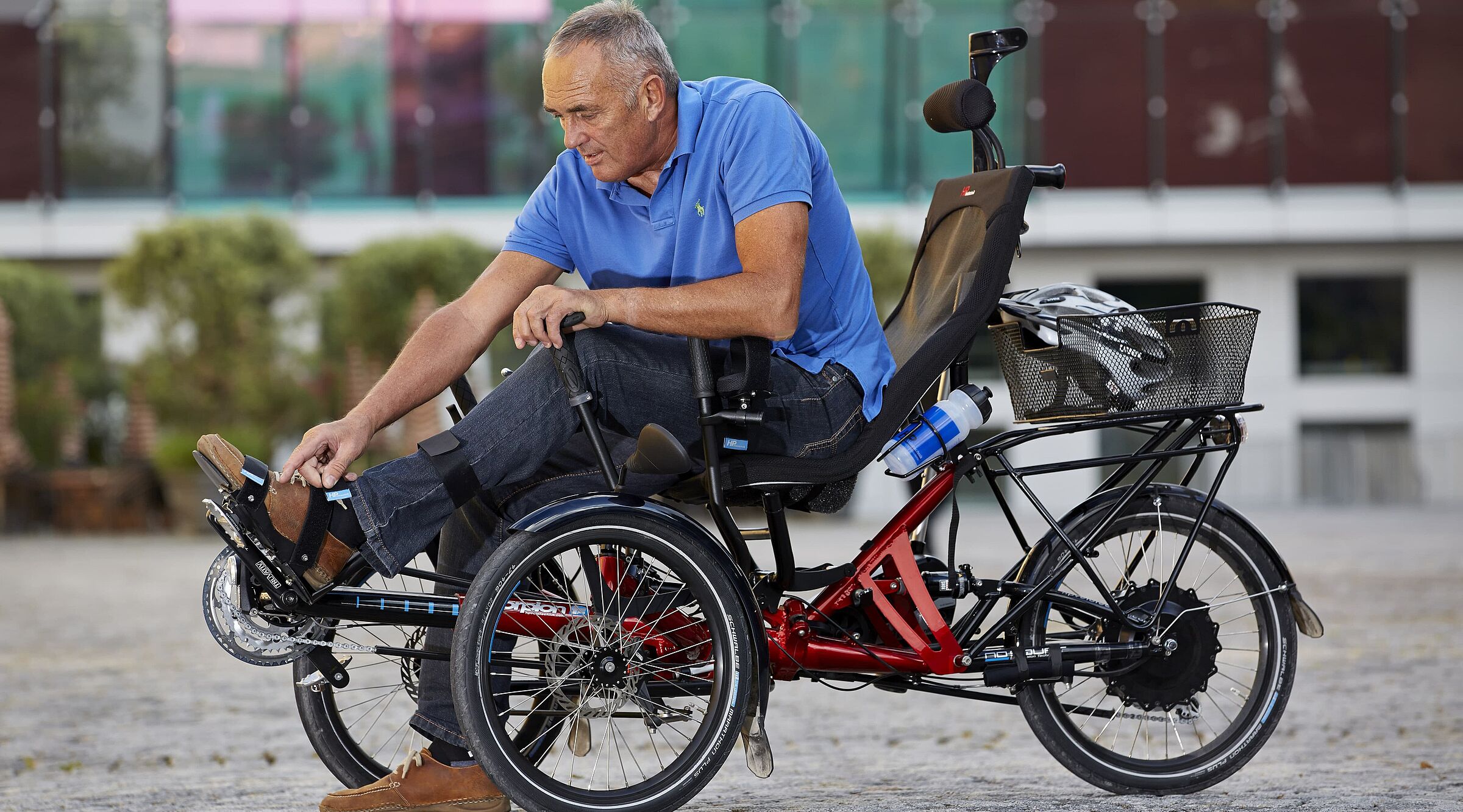 Ein Mann in blauem Poloshirt sitzt auf einem roten dreirädrigen Elektrofahrrad mit schwarzer Rückenlehne und Korb. Das Spezialrad steht auf einem gepflasterten Platz vor einem modernen Gebäude mit grünen Fenstern.