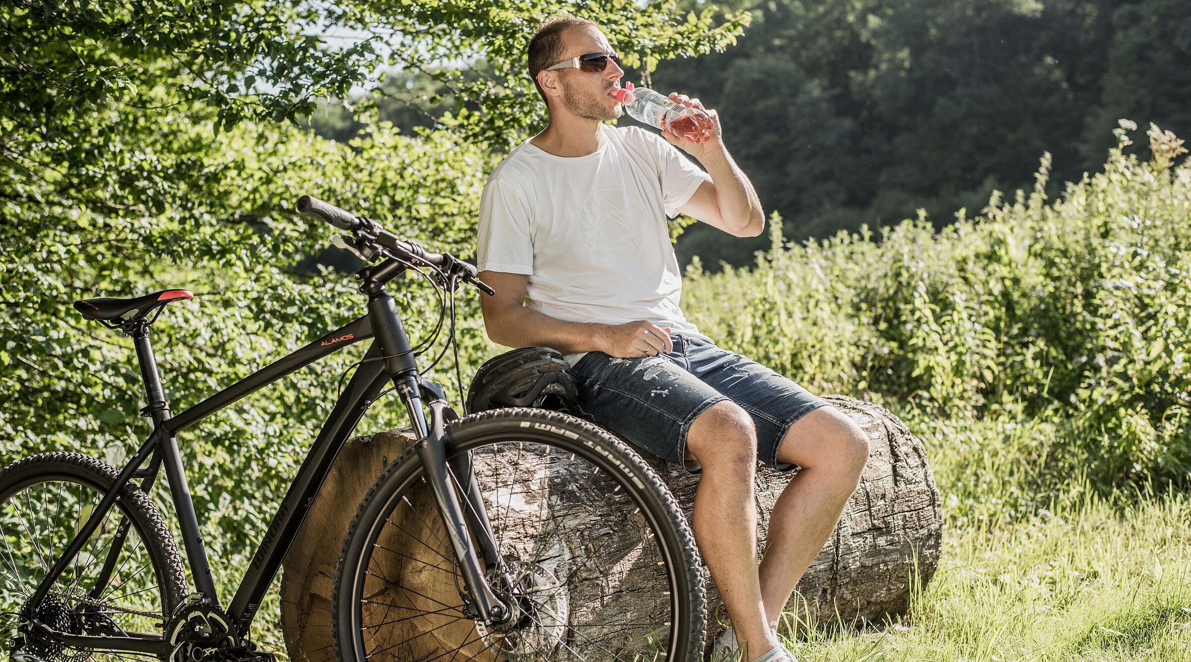 Radfahrer mit Sonnenbrille trinkt Wasser aus einer Flasche während einer Pause auf einem Baumstamm. Sein schwarzes Mountainbike steht daneben.