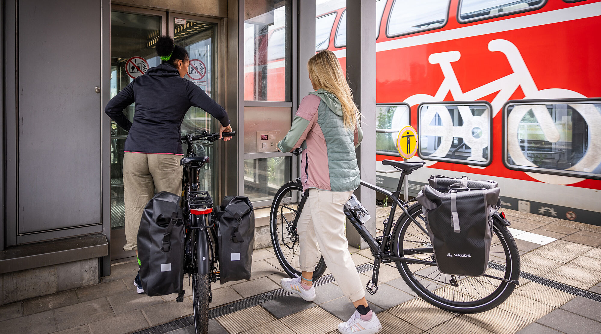 Zwei Frauen schieben zwei Fahrräder in einen Aufzug am Bahnhof. Im Hinterung ein Regionalzug mit Fahrradsymbol.