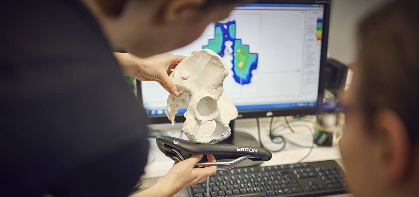 Eine Person hält ein Modell eines Beckens auf einen Fahrradsattel. Das Foto veranschaulicht die Analyse der Ergonomie zwischen menschlicher Anatomie und Fahrradsitz.