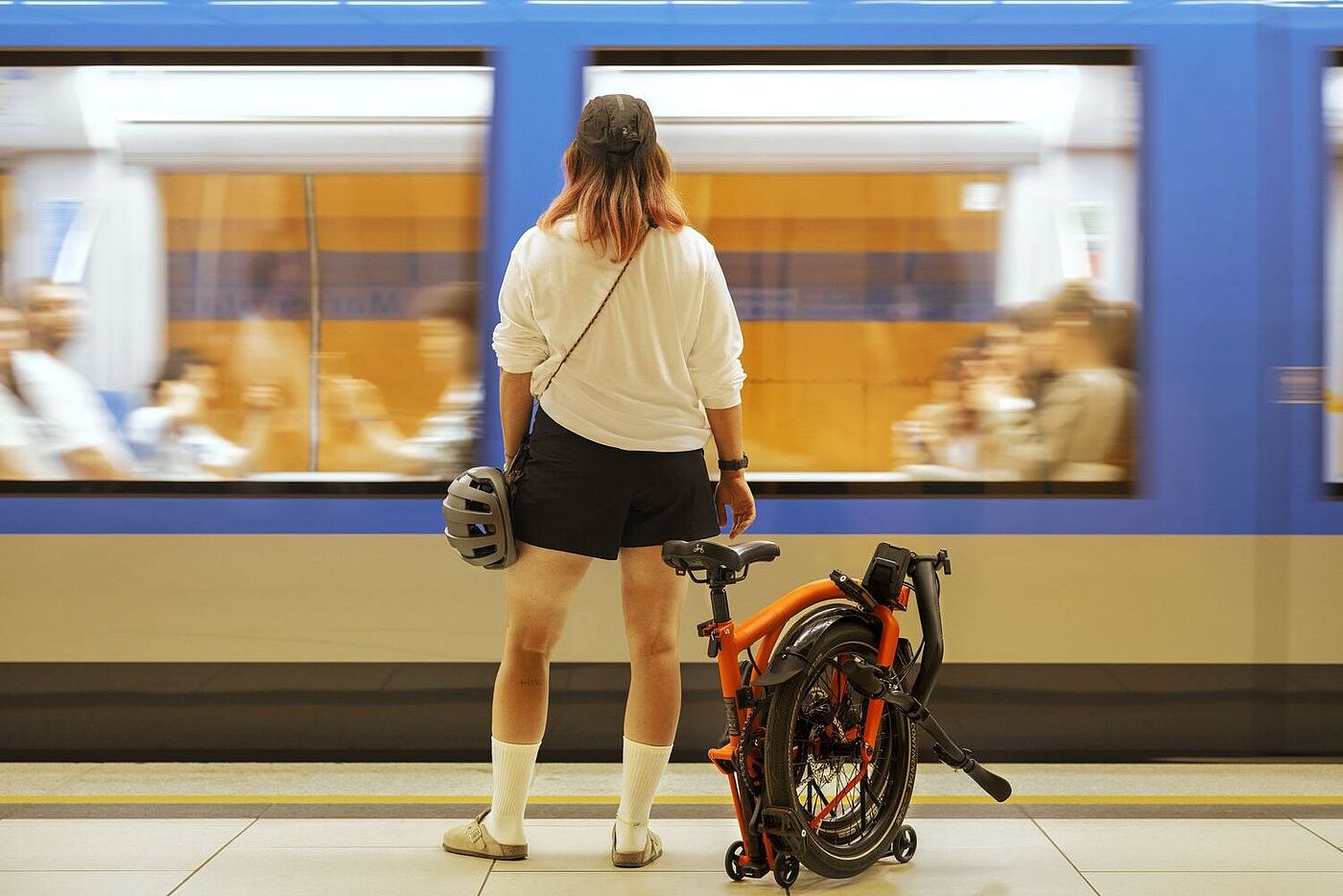 Eine Frau steht am Bahnsteig mit einem gefalteten Brompton G-Line neben sich. 